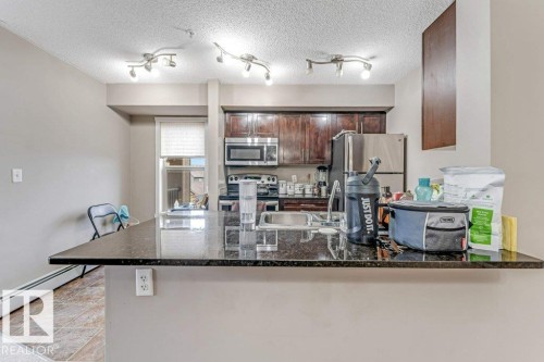 Kitchen featuring rich wood-finish cabinetry, stainless steel appliances, a dark granite-style countertop peninsula, and track lighting fixtures - 225 111 Watt Common, Edmonton, AB 
