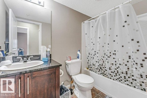 Bathroom featuring a dark wood-finish vanity with a built-in sink and dark countertop, a toilet, a full-size bathtub, and a tiled shower surround - 225 111 Watt Common, Edmonton, AB 