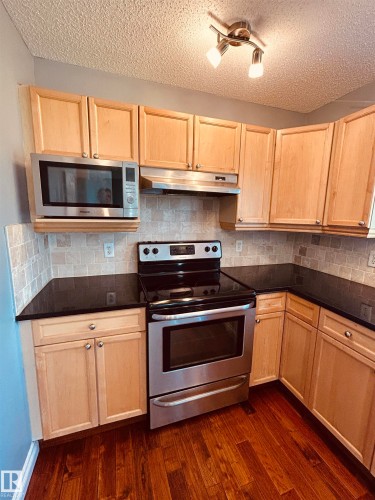 Kitchen featuring natural wood-finish cabinetry, black countertops, and a stainless steel appliance suite - 89 12050 17 Avenue, Edmonton, AB 