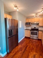 Kitchen featuring wood-finish flooring, stainless steel appliances, light wood cabinetry, dark countertops, and a neutral tile backsplash - 
