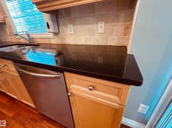 Kitchen featuring light wood cabinetry with round silver hardware, dark countertops, a stainless steel dishwasher, a stainless steel sink with a gooseneck faucet, and a tiled backsplash - 