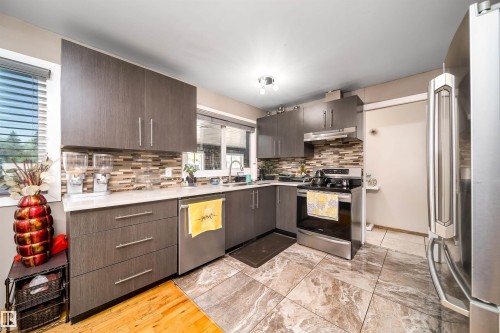 Modern kitchen featuring wood-grain cabinetry, stainless steel appliances, a mosaic tile backsplash, and stone-look tile flooring - 14808 96 Street, Edmonton, AB 