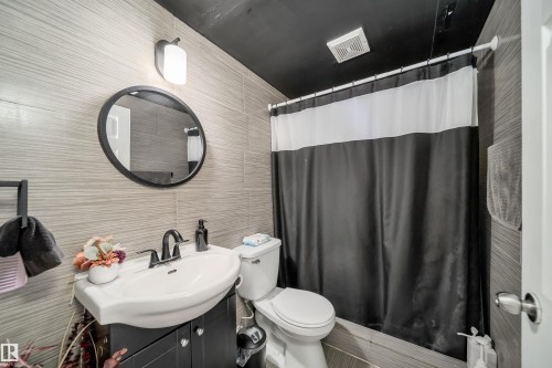 Bathroom featuring textured wall tiling, a black vanity with a white basin, a circular black-framed mirror, and a black ceiling - 14808 96 Street, Edmonton, AB 