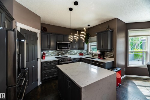 Contemporary kitchen featuring a large central island with a light-colored countertop, dark cabinetry, stainless steel appliances, a multi-tone backsplash, and modern pendant lighting - 704 Wildwood Point(E), Edmonton, AB 