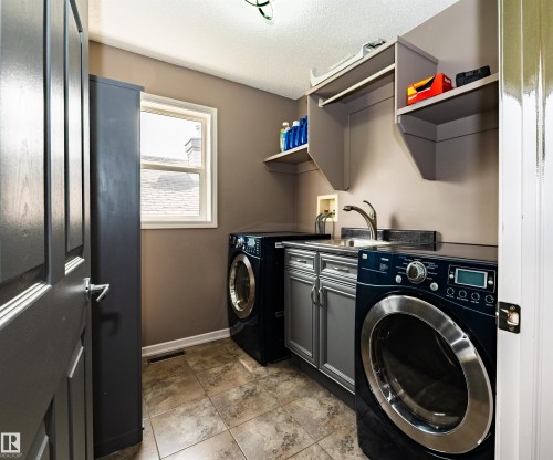 Dedicated laundry area featuring a utility sink, under-sink cabinetry, open shelving, window, and tile flooring - 704 Wildwood Point(E), Edmonton, AB 