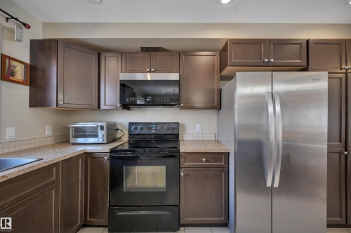 Contemporary kitchen featuring dark wood cabinetry, a stainless steel refrigerator, black range, and an integrated stainless steel microwave - 29 1150 Windermere Way, Edmonton, AB 