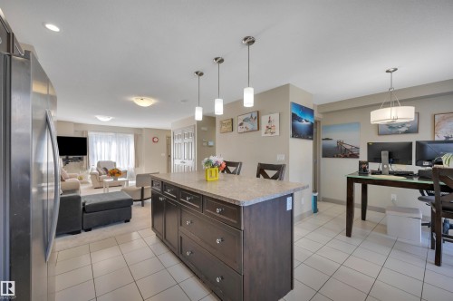 Open-concept living area featuring a kitchen island with pendant lighting, a stainless steel refrigerator, tile flooring, and carpeted living room space - 29 1150 Windermere Way, Edmonton, AB 