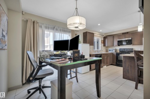 Open-concept area featuring white tile flooring, a large window with sheer and blackout curtains, an overhead drum pendant light, and a kitchen with dark cabinetry and black appliances - 29 1150 Windermere Way, Edmonton, AB 