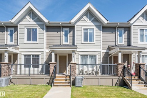 Modern townhome exterior featuring light gray horizontal siding, white trim, prominent gables, a front porch with black metal railings, and stone-clad pillars - 29 1150 Windermere Way, Edmonton, AB 