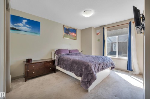 Carpeted bedroom featuring a large window with white trim, a flush-mount ceiling light fixture, neutral wall paint, and a built-in television mount - 29 1150 Windermere Way, Edmonton, AB 