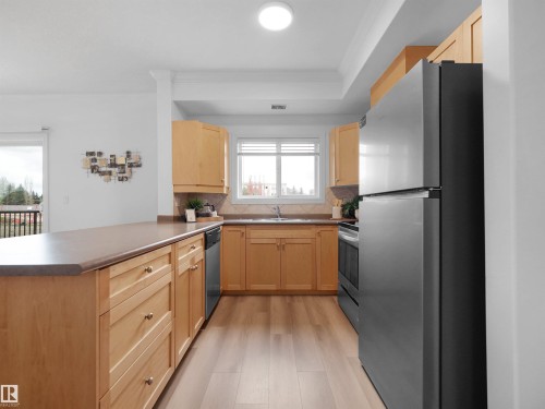 Galley kitchen featuring wood-finish flooring, light wood cabinetry, dark countertops, and a window above the sink - 104 14612 125 Street, Edmonton, AB 