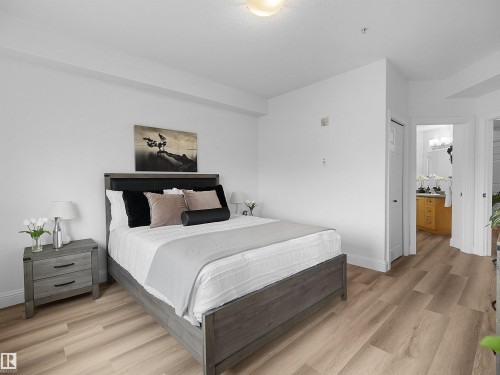 Spacious bedroom featuring wood-finish flooring, a contemporary bed frame with an upholstered headboard, and a white color palette - 104 14612 125 Street, Edmonton, AB 