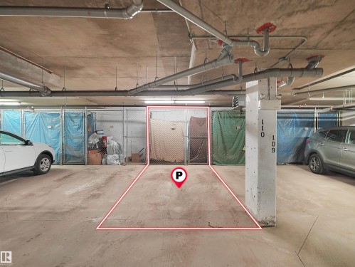 Designated parking space within a concrete garage structure, featuring exposed overhead piping, concrete support columns, and chain-link storage units - 104 14612 125 Street, Edmonton, AB 