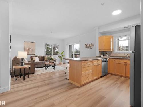 Open-concept living space featuring wood-finish flooring, a kitchen island with seating, natural wood cabinetry, and a sliding glass door leading to an outdoor area - 104 14612 125 Street, Edmonton, AB 