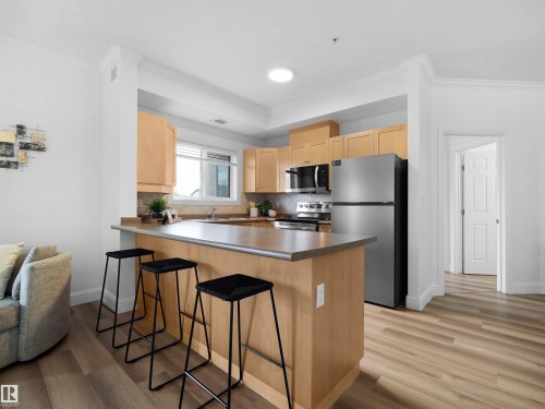 Kitchen featuring light wood cabinetry, stainless steel appliances, a breakfast bar, and wood-finish flooring - 104 14612 125 Street, Edmonton, AB 