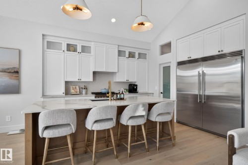 Modern kitchen featuring a large central island with seating, a built-in stainless steel refrigerator, white cabinetry with glass accents, light wood-finish flooring, and vaulted ceilings - 14 Jane Point(E), St. Albert, AB 