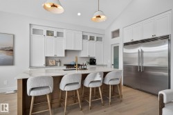 Modern kitchen featuring a large central island with seating, a built-in stainless steel refrigerator, white cabinetry with glass accents, light wood-finish flooring, and vaulted ceilings - 