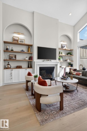 Bright living area featuring a tall fireplace with a white mantel, arched built-in shelving with accent lighting, white cabinetry, and wood-finish flooring - 14 Jane Point(E), St. Albert, AB 