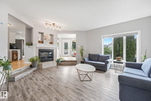 Living area featuring wood-finish flooring, a corner fireplace with a tile surround and hearth, and a large picture window - 98 Bridgeport Wynd, Leduc, AB 