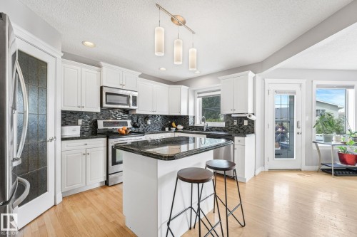 Modern kitchen featuring light wood-finish flooring, white cabinetry, dark stone countertops, and a dark mosaic tile backsplash - 98 Bridgeport Wynd, Leduc, AB 