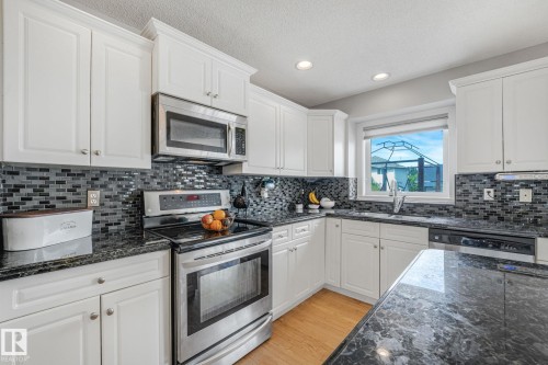 Kitchen featuring white cabinetry with crown molding, stainless steel appliances, dark stone countertops, and a mosaic tile backsplash - 98 Bridgeport Wynd, Leduc, AB 