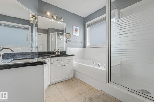 Bathroom featuring a dual vanity with dark countertops, white cabinetry, a built-in tub, and a glass-enclosed shower - 98 Bridgeport Wynd, Leduc, AB 