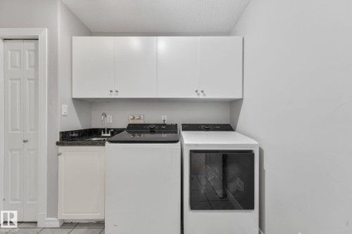 Dedicated laundry area featuring a utility sink with a stainless steel faucet, dark stone-finish countertop, and upper white cabinetry for storage - 98 Bridgeport Wynd, Leduc, AB 