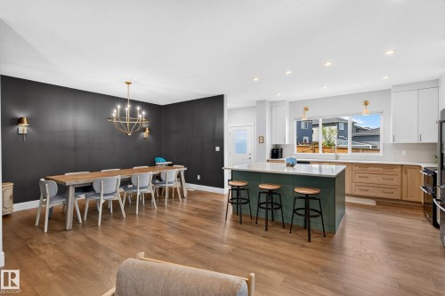 Open-concept kitchen and dining area featuring wood-finish flooring, a kitchen island with a light-colored countertop, white upper cabinetry, natural wood-finish lower cabinetry, and a gold-finish chandelier - 30 Rosewood Way, St. Albert, AB 