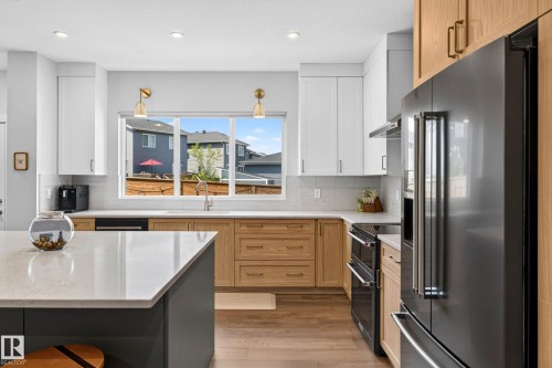 Kitchen featuring a large central island with a light-toned countertop, a window-mounted sink, two-tone cabinetry with light wood-finish lower cabinets and white upper cabinets, and dark stainless steel appliances - 30 Rosewood Way, St. Albert, AB 