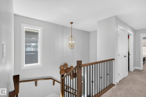 Upper-level landing featuring a wood-finish handrail with wrought iron spindles, a prominent window with white trim, a decorative pendant light fixture, and light-toned carpeting - 30 Rosewood Way, St. Albert, AB 