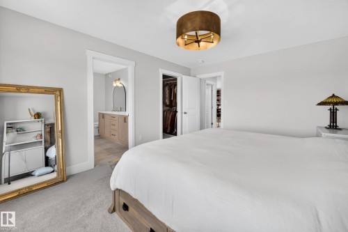 Carpeted bedroom featuring a large white bed, a drum pendant light fixture, and an en-suite bathroom with a wood-finish vanity and an arched mirror - 30 Rosewood Way, St. Albert, AB 
