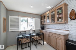Kitchen featuring light wood cabinetry with glass-front upper cabinets, a white brick-finish accent wall, and a window with white louvered shutters - 