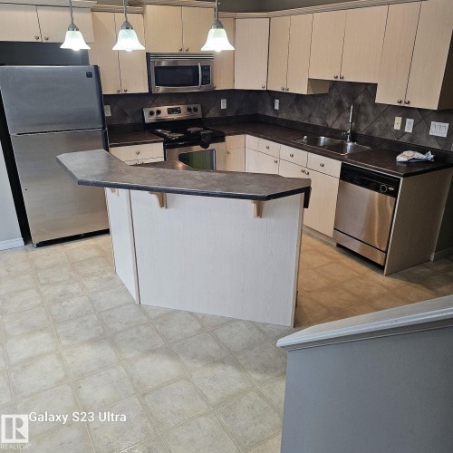 Well-appointed kitchen featuring light-toned cabinetry, dark countertops, and a full suite of stainless steel appliances including a refrigerator, range, microwave, and dishwasher - 99 13825 155 Avenue, Edmonton, AB 