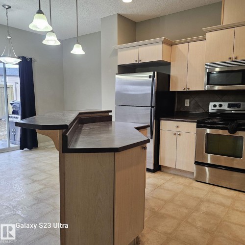 Kitchen featuring an integrated breakfast bar with a dark countertop, light wood-finish cabinetry, stainless steel appliances, and a tiled backsplash - 99 13825 155 Avenue, Edmonton, AB 