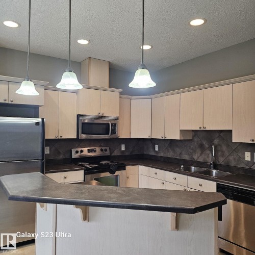 Kitchen featuring a peninsula breakfast bar, stainless steel appliances, light wood-finish cabinetry, dark countertops, and a tiled backsplash - 99 13825 155 Avenue, Edmonton, AB 