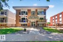 Brick facade building featuring multiple private balconies with black railings, a central entrance with an awning, manicured lawn areas, and a concrete pathway leading to the entrance - 33 10230 122 Street, Edmonton, AB 
