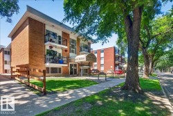 Brick multi-story building featuring individual balconies with black metal railings, a white awning over the main entrance, and mature trees lining the street - 