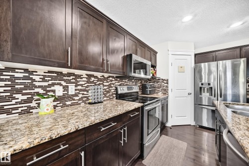 Kitchen featuring dark wood cabinetry, light-toned stone-finish countertops, a mosaic tile backsplash, and stainless steel appliances including a refrigerator, range, and microwave - 3433 Goodridge Link Link, Edmonton, AB 