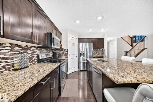 Well-appointed kitchen featuring dark wood cabinetry, stainless steel appliances, a mosaic tile backsplash, and extensive granite countertops including a large island with integrated sink - 3433 Goodridge Link Link, Edmonton, AB 