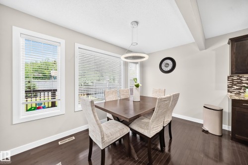 Dining area featuring wood-finish flooring, two large windows with blinds, a contemporary pendant light fixture, and a partial view of kitchen cabinetry with a mosaic tile backsplash - 3433 Goodridge Link Link, Edmonton, AB 