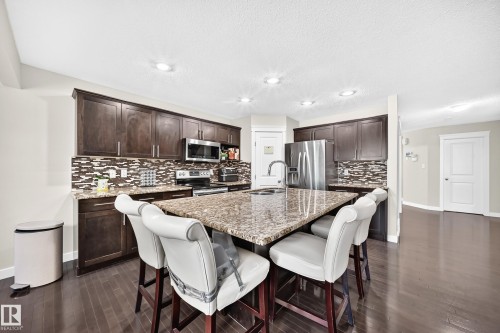 Open-concept kitchen featuring dark wood cabinetry, granite countertops, a mosaic tile backsplash, stainless steel appliances, and wood-finish flooring - 3433 Goodridge Link Link, Edmonton, AB 