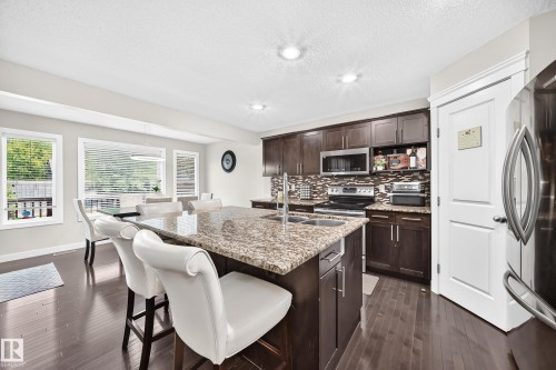 Kitchen featuring a large island with a stone countertop and double sink, dark wood cabinetry, stainless steel appliances, and a mosaic tile backsplash - 3433 Goodridge Link Link, Edmonton, AB 