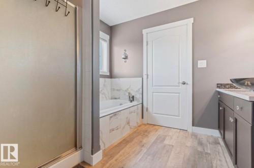 Bathroom featuring a built-in tub with marble-look tile surround, a glass-enclosed shower, wood-finish flooring, a vanity with a vessel sink, and a white interior door - 1007 Macewan Cl, Edmonton, AB 