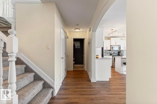 Open concept interior featuring wood-finish flooring, a carpeted staircase with white balusters, an arched doorway, white kitchen cabinetry, and stainless steel appliances - 1007 Macewan Cl, Edmonton, AB 
