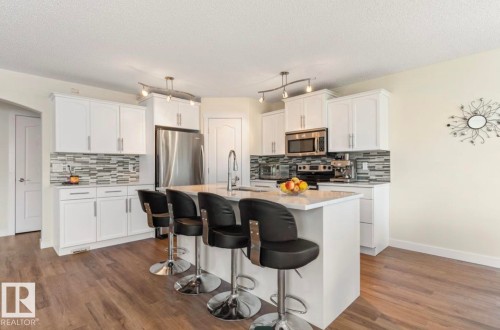 Modern kitchen featuring white cabinetry, stainless steel appliances, a mosaic tile backsplash, a central island with a sink, and wood-finish flooring - 1007 Macewan Cl, Edmonton, AB 