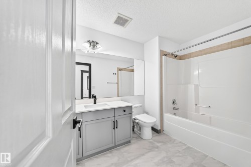 Bathroom featuring a white tub and shower combination with tan tile surround, a gray vanity with an integrated sink and black faucet, a large wall-mounted mirror, and marble-look floor tiles - 1549 37C Avenue, Edmonton, AB 