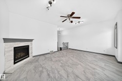 Spacious room featuring light-toned wood-finish flooring, a contemporary fireplace with a stone-look surround and dark mantel, a ceiling fan, track lighting, and white painted walls - 