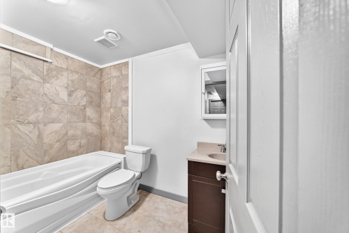 Bathroom featuring a tub with stone-look tile surround, a white toilet, and a vanity with an undermount sink and stone countertop - 1549 37C Avenue, Edmonton, AB 