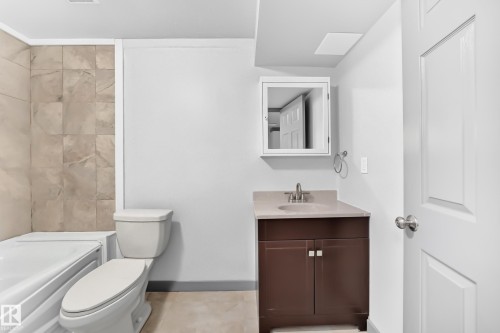 Bathroom featuring a dark wood-finish vanity with a light countertop and undermount sink, a mirrored medicine cabinet, a white toilet, and a white bathtub with light-toned tiled surrounds - 1549 37C Avenue, Edmonton, AB 