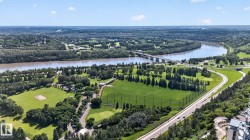 Expansive aerial perspective showcasing a wide river, lush green parklands, a multi-lane roadway, a prominent bridge, and densely wooded areas - 